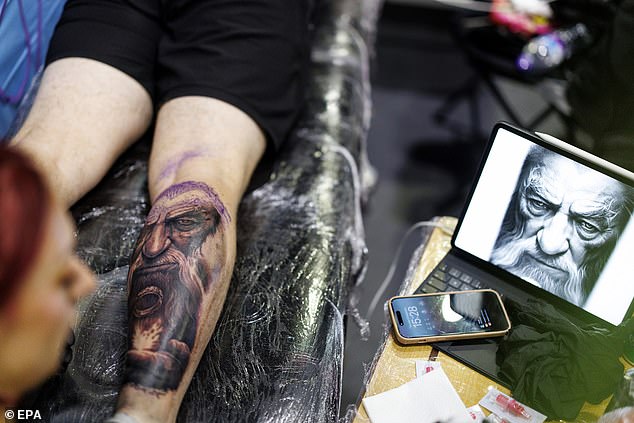 A tattoo artist carefully looks at a reference picture as she draws out a design on a customer's leg