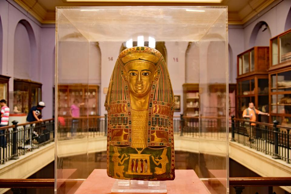 Gold Egyptian funeral casket mask in a display case.