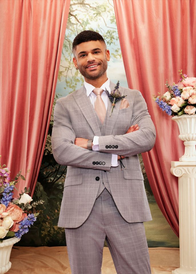 Keye in a light grey plaid suit and peach tie, with a boutonniere, smiling with arms crossed against a backdrop of peach curtains, painted trees, and floral arrangements.