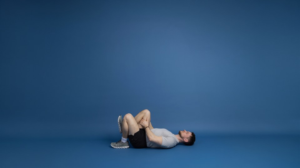 A man in a light grey t-shirt and black shorts lies on his back with his knees bent and pulled towards his chest, holding his knees with his hands.