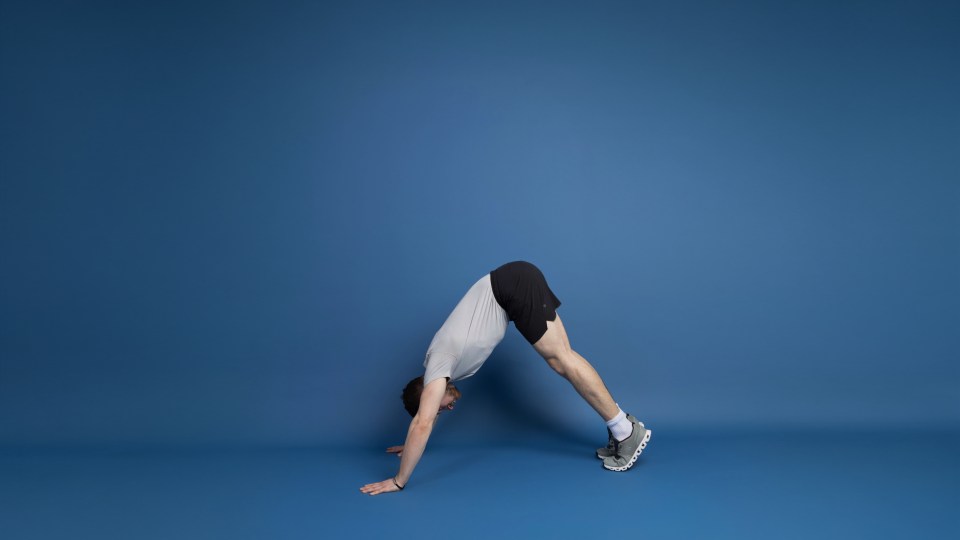A man in a light gray shirt and black shorts demonstrating a downward-facing dog yoga pose.