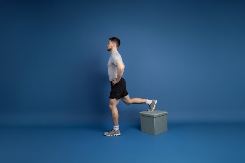 Man in athletic wear demonstrating a Bulgarian split squat with his back foot on a gray box.