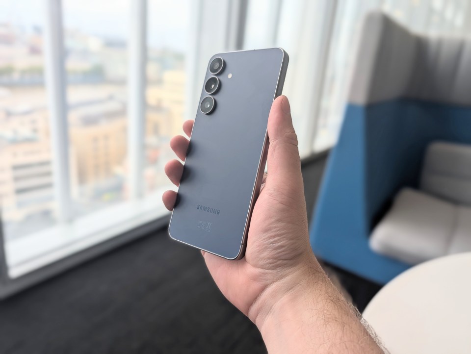 A person's hand holding a gray Samsung phone with three camera lenses on the back.