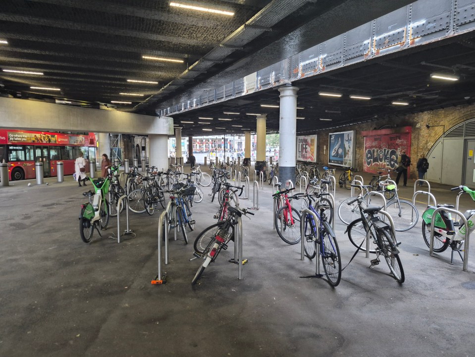 Numerous bicycles parked in racks under a dark overpass, with advertisements on the walls and a red double-decker bus in the background.