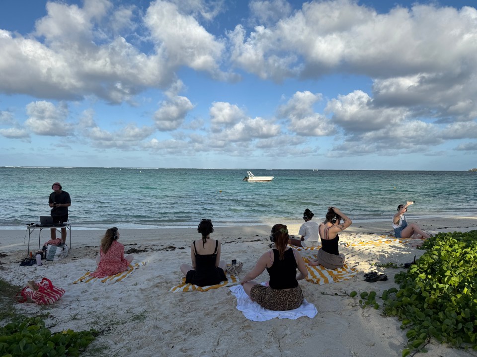 A group of people wearing headphones sit on a sandy beach looking out at the ocean under a cloudy sky.