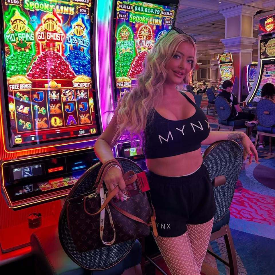 Linzee Ryder in a casino wearing a black sports bra and shorts with fishnet stockings, next to a slot machine.