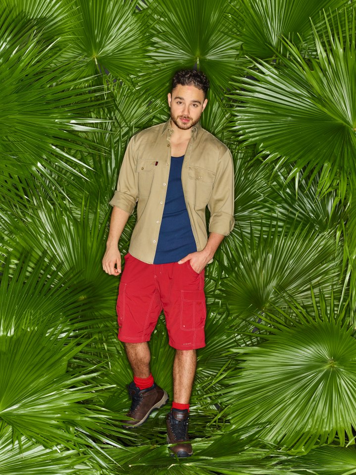Adam Thomas, wearing a tan shirt, blue undershirt, red shorts, and brown hiking boots, stands among large palm leaves.