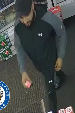 Mohammed Maneer Khan, wanted for murder, wearing a black and gray jacket, black pants, and a black baseball cap, holding a small object.