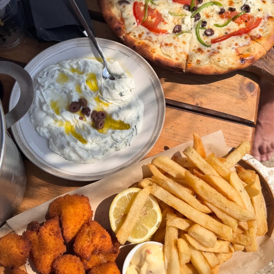 A wooden table with a plate of tzatziki dip with olives and olive oil, a pizza with tomatoes, bell peppers, olives, and cheese, and a tray of fried food including french fries, lemon, and a dip.