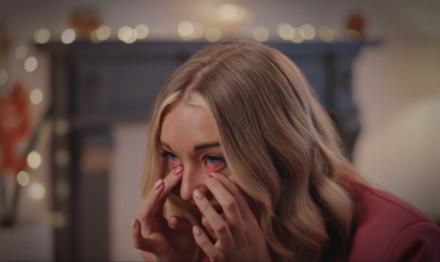 A blonde woman with blue eyes, red eyelids, and red around her eyes, pushes up her eyelids with her index fingers.