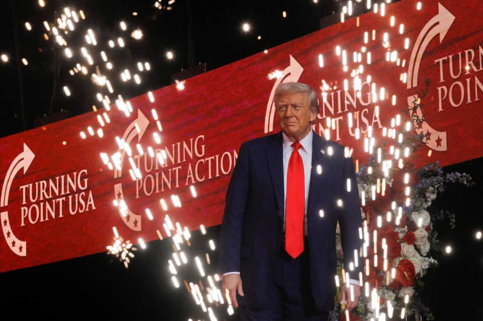 U.S. President Donald Trump stands onstage in front of a red banner that reads "TURNING POINT USA" and "TURNING POINT ACTION" with fireworks around him.
