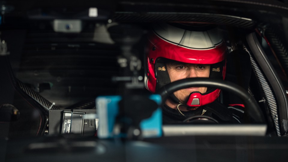 A driver in a red and white racing helmet behind the wheel of a performance car.