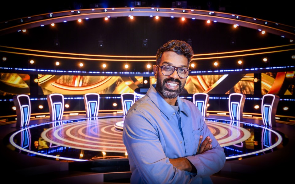 Comedian Romesh Ranganathan stands smiling in a blue shirt with his arms crossed on a game show set.