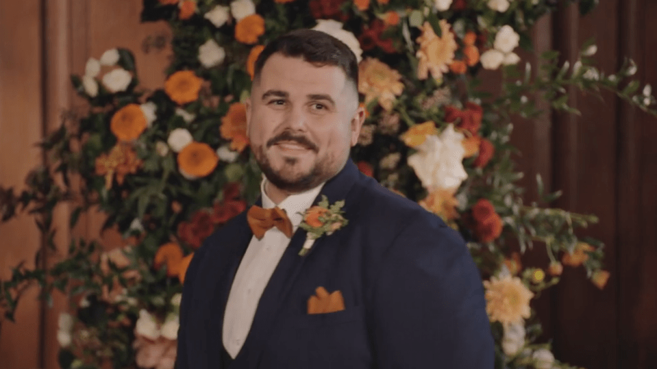 A man in a navy suit with an orange bowtie and boutonnière smiling in front of a floral background.