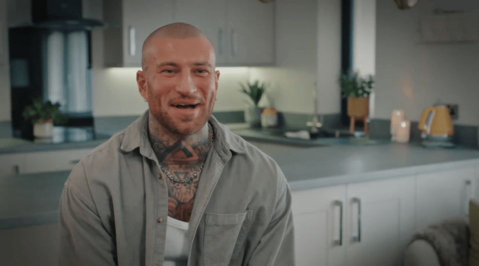 A bald man with neck tattoos and a chain necklace smiles at the camera in a kitchen.