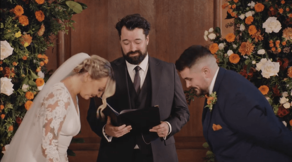 A wedding ceremony with a bride, groom, and officiant standing before an altar adorned with orange and white flowers.