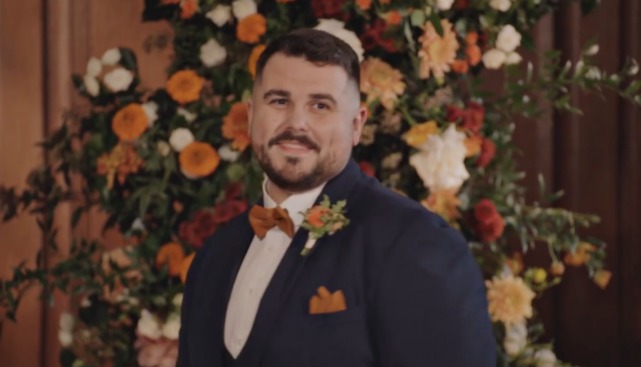 Groom in a navy suit with an orange bowtie and boutonniere, smiling in front of a floral arrangement.