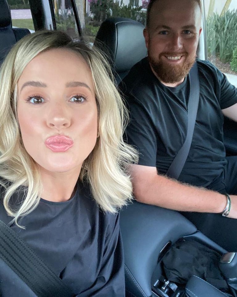 A blonde woman making a kiss face and a bearded man smiling, both in a car.