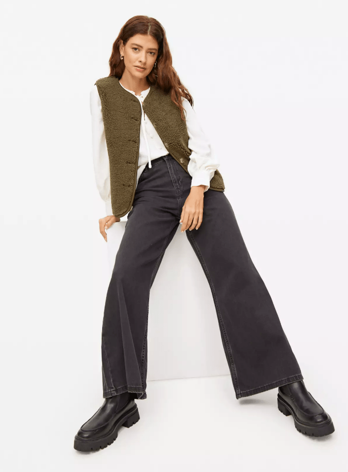 A woman wearing a white blouse, a green textured vest, wide-leg gray jeans, and black chunky boots, posing with one leg bent.