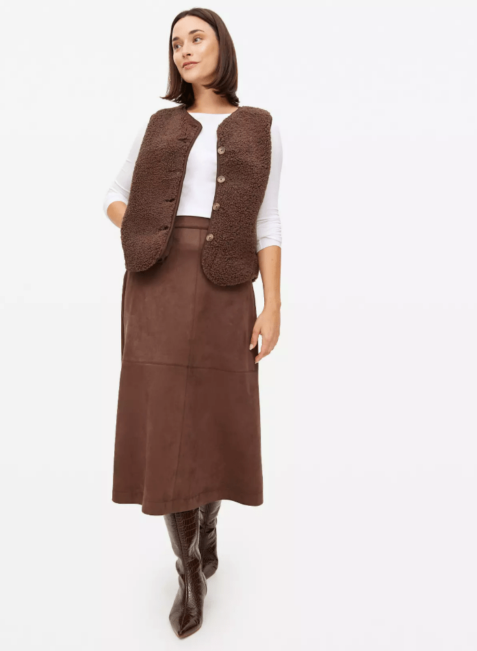 A woman models a long-sleeve white shirt with a brown vest, brown skirt, and brown boots.