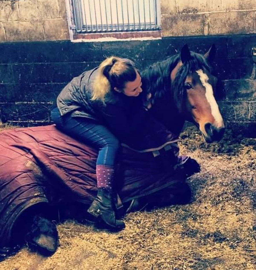A woman in a blue jacket and jeans hugging a brown and white horse that is lying down.