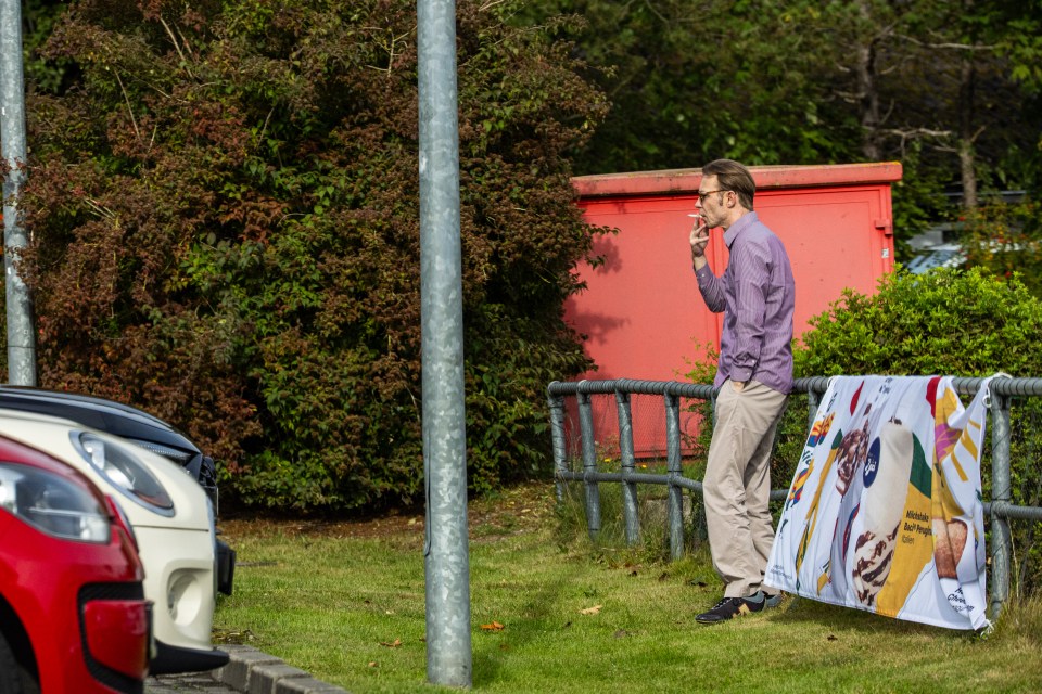 Christian Brueckner smoking next to parked cars and a red building.