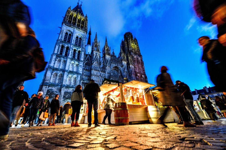 People gather at the Christmas Market in Rouen's Place de la Cathedrale square, with the cathedral looming in the background under a dark blue sky.