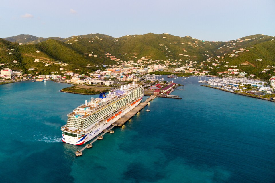 Cruise ship Arvia docked at a port in the Caribbean, with a town and green hills in the background.