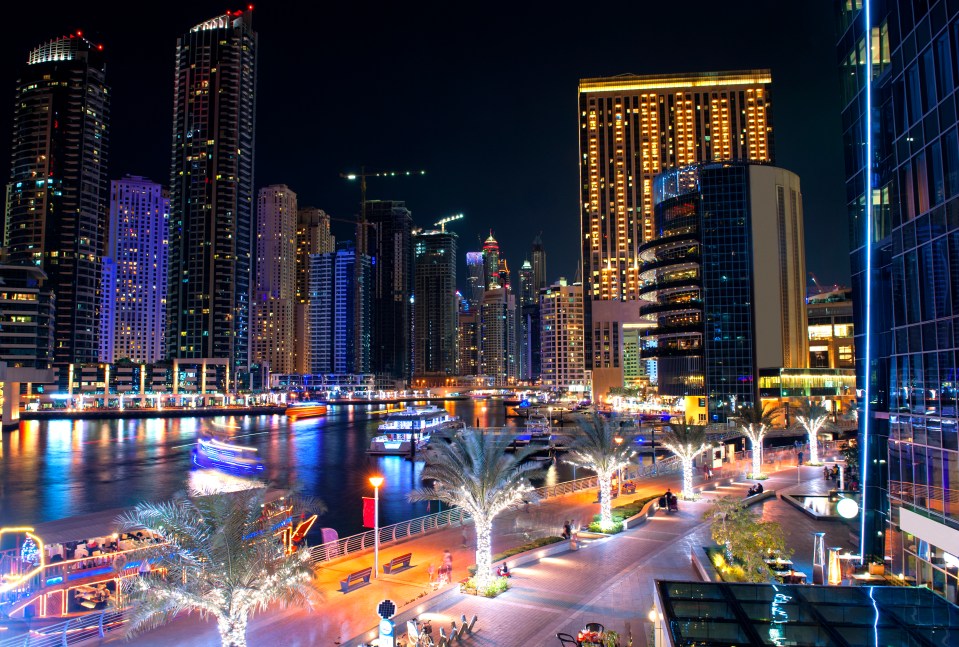 Dubai Marina at night with brightly lit skyscrapers, boats, and palm trees with Christmas lights.