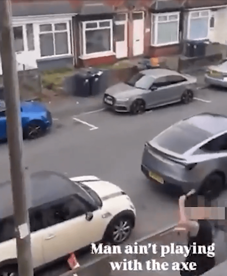 A person holding a blurred object, possibly an axe, standing near parked cars on a residential street.