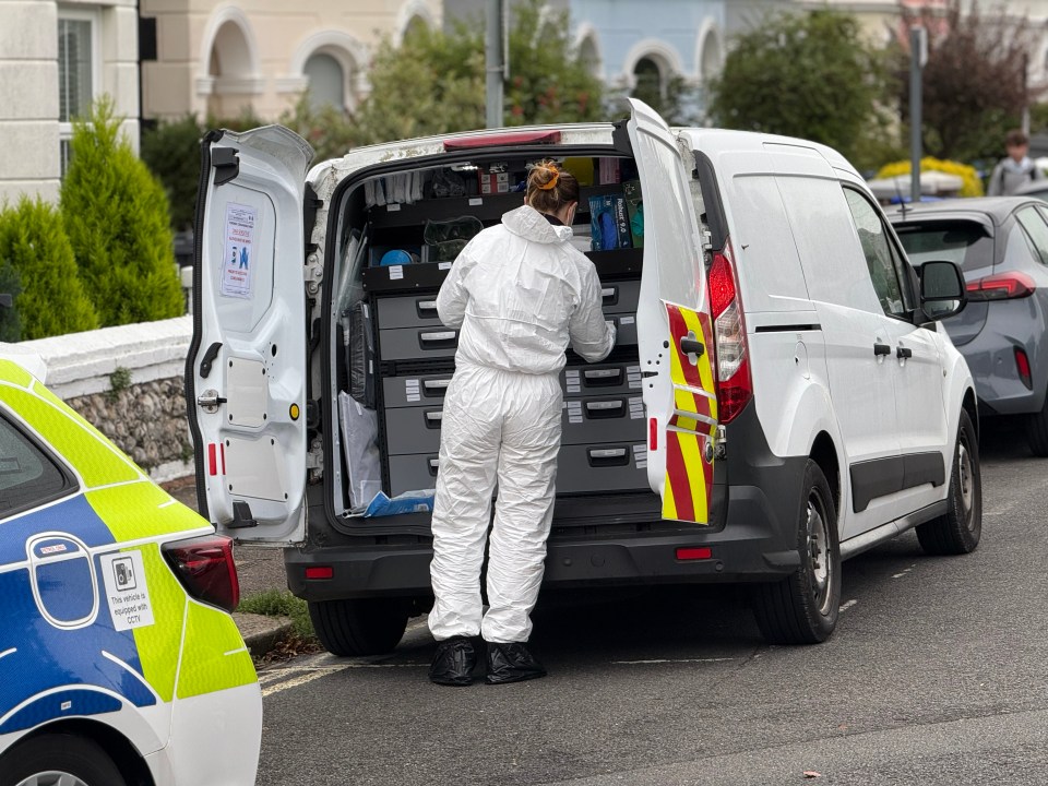 A person in a white forensic suit and black boot covers stands at the back of a white van with its rear doors open, organizing items. A police vehicle is partially visible on the left.