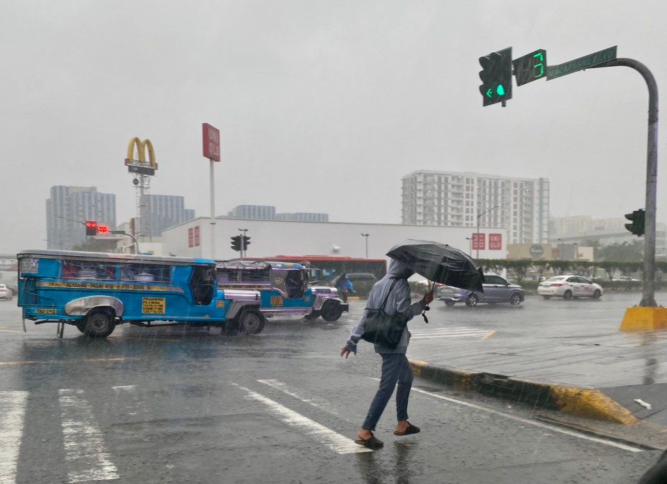 A Filipino walks through heavy rain in Manila with an umbrella and a jeepney in the background.
