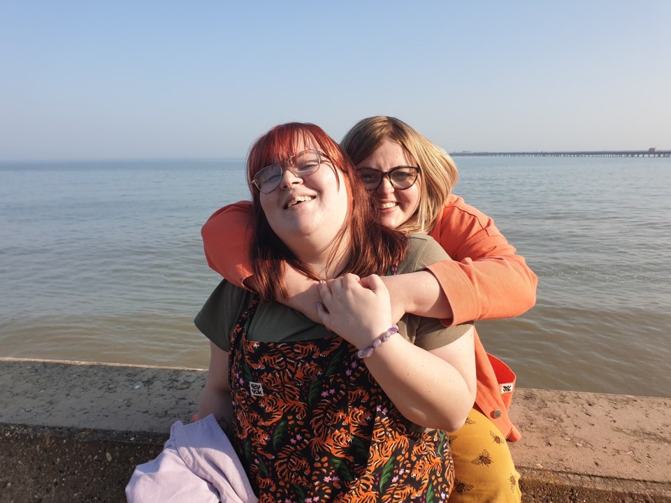 Zoe Nunn (left) and her sister Lily Nunn (right) smiling at the camera, with Zoe's arms around Lily, by a body of water with a pier in the distance.