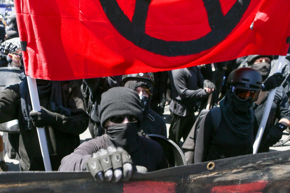 Antifa members and counter-protesters gather during a right-wing "No-To-Marxism" rally, with one person in the foreground holding a pole supporting a red flag with a black symbol.