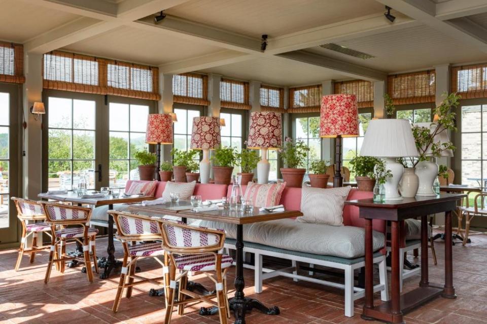 Restaurant interior with multiple dining tables, chairs, and benches, potted plants, and large windows with outdoor views.