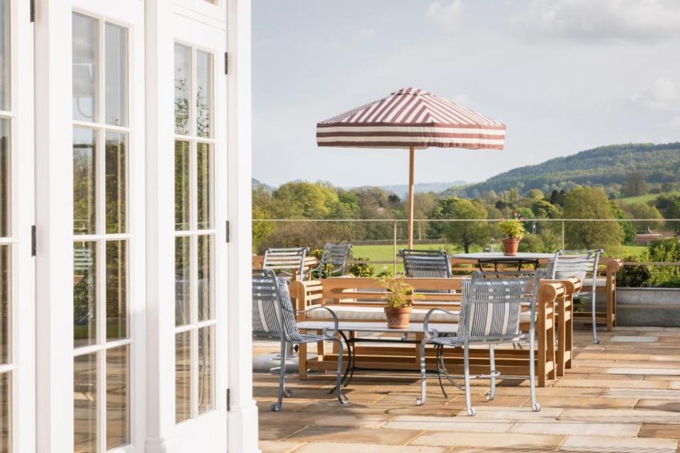 Outdoor patio with tables, chairs, and a striped umbrella, overlooking a green valley.