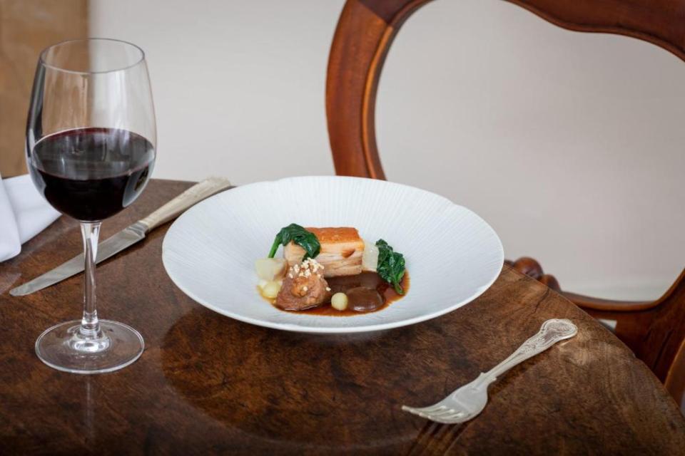 A finely plated dish of pork belly with greens and sauce, accompanied by a glass of red wine, a knife, and a fork, on a wooden table.