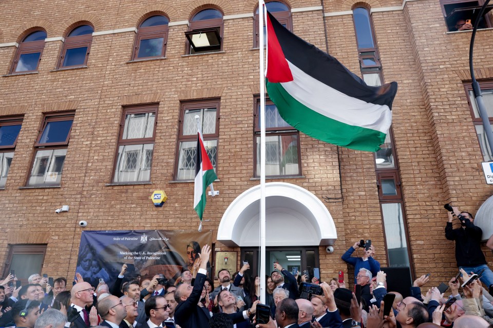 The Palestinian flag is raised at the Palestinian mission in London, watched by a crowd of people.