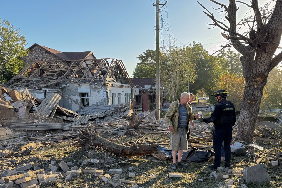A psychologist aiding a man at a Russian airstrike site with a body bag and a destroyed house in the background.