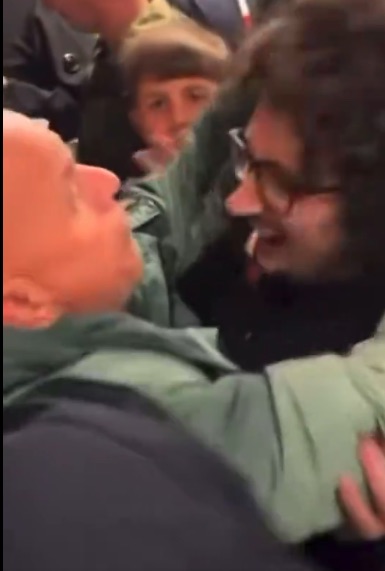 A close-up of a person with curly hair and glasses leaning in to speak to an older man.
