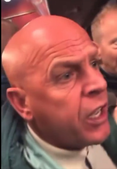 Close-up of a bald man with light skin, wrinkles on his forehead, and an open mouth as if speaking loudly.