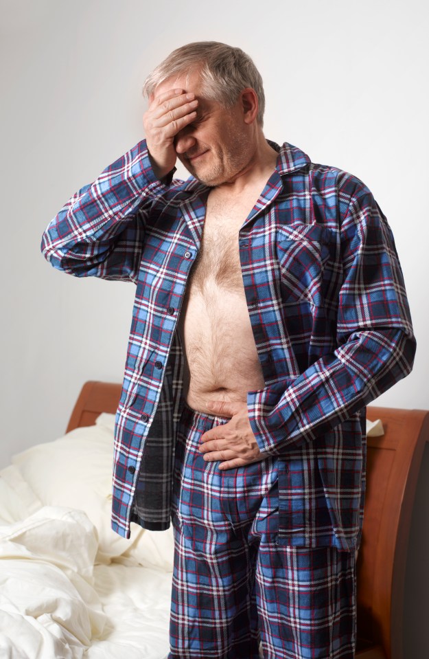 A mature man in blue plaid pajamas clutching his stomach with one hand and forehead with the other, indicating stomach pain.