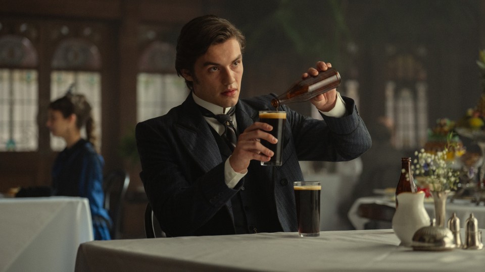 Louis Partridge pouring dark beer from a bottle into a glass.