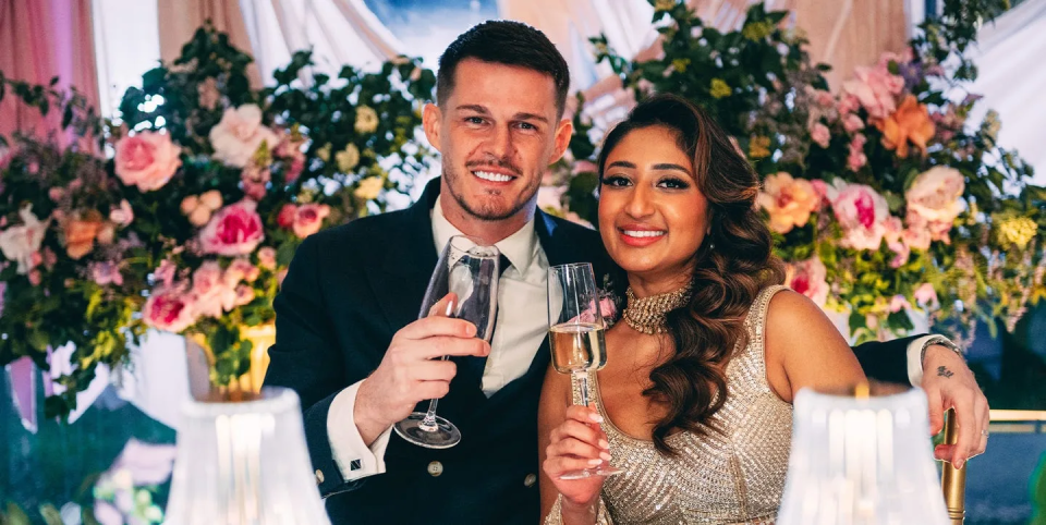 A man and woman smiling and holding champagne glasses in front of floral arrangements.