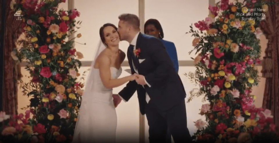 A groom kissing his bride during their wedding ceremony, with floral arches on either side.