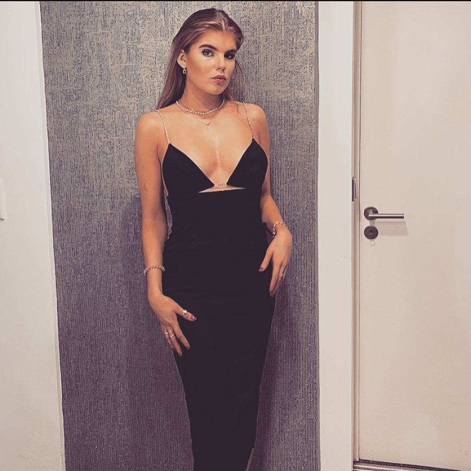 A woman in a black dress with spaghetti straps and cutouts at the bust, standing against a textured wall next to a white door.