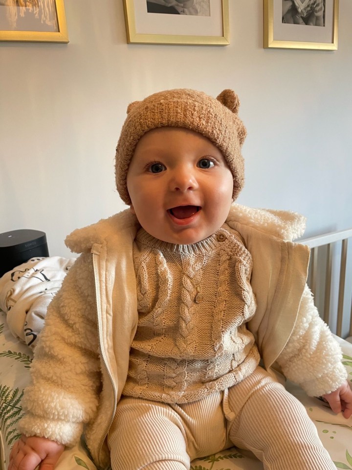 Tommy Francis Kneebone, a 7-month-old baby, smiles while wearing a cream-colored outfit and a brown bear-eared hat.
