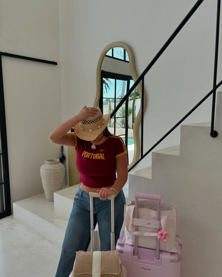 Alanna Pow, wearing a straw hat and a red Portugal t-shirt, standing with her luggage.