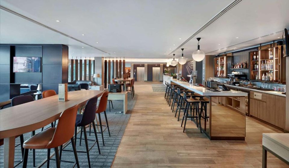 A spacious hotel lounge area with a long wooden table and chairs, a bar with high stools, and two elevators in the background.