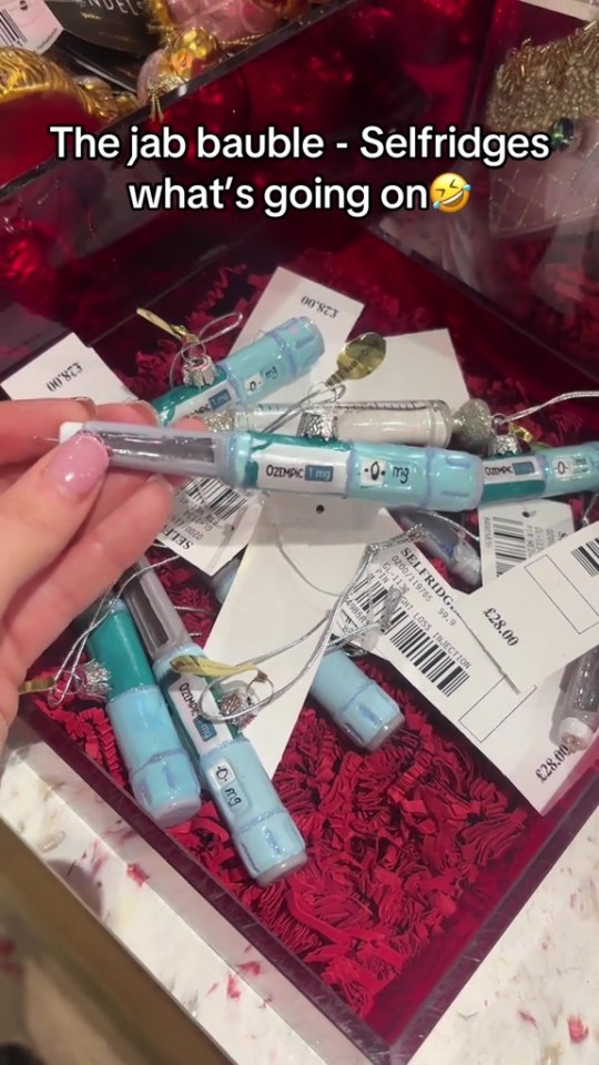 A hand holding an Ozempic pen-shaped Christmas bauble from a box filled with similar baubles and price tags.